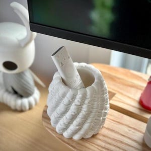 May include: A white, textured, cylindrical holder with a spiraling, ribbed pattern containing a slim, white remote control. The holder has a matte finish and a hollow center, providing a stable base for the remote to stand upright.