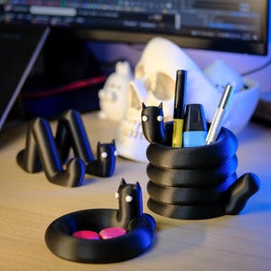 May include: Black 3D-printed desk organizers in the shape of a snake with cat-like features. The set includes a pen holder, a small dish with pink objects, and a decorative snake-shaped piece. The pen holder contains pens and markers.