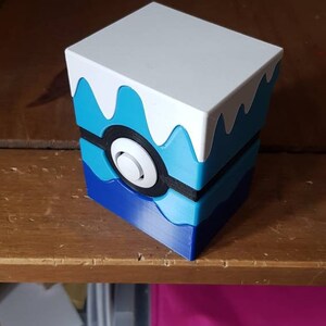May include: A cube-shaped storage box with a white top and a blue and white design resembling a stylized wave. The box features a black and white circular design on the front, reminiscent of a Pokeball.