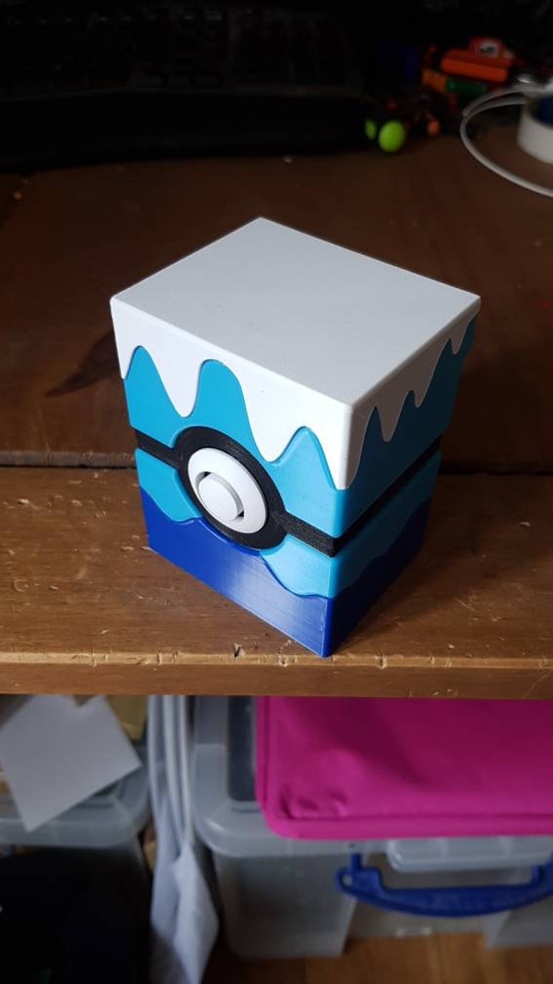 May include: A cube-shaped storage box with a white top and a blue and white design resembling a stylized wave. The box features a black and white circular design on the front, reminiscent of a Pokeball.