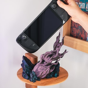 Può includere: Console di gioco portatile nera sopra un ripiano in legno con una scultura decorativa viola e blu. Il ripiano è montato su una parete bianca, con una libreria in legno sullo sfondo.