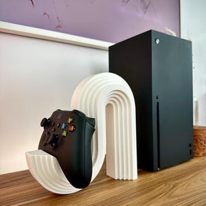 Peut inclure: Une manette Xbox noire repose dans un support blanc en forme d'arche avec un design nervuré. Une console Xbox Series X noire est debout à côté du support. Les articles sont sur une surface en bois, avec de l'art abstrait en arrière-plan.
