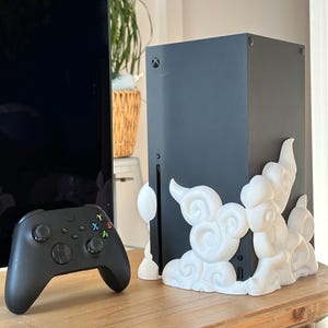 May include: Black Xbox Series X console with a white cloud-shaped decorative stand. A black Xbox controller is on the left. The console sits on a wooden surface.