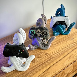 Könnte beinhalten: Drei Tintenfisch-Controller-Halter in Weiß, Grau und Blau. Jeder Halter hält einen anderen Gamecontroller: einen schwarzen Xbox-Controller, einen grauen Controller und einen weiß-schwarzen PlayStation-Controller. Die Halter stehen auf einer Holzoberfläche.