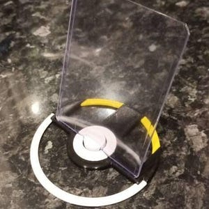 Puede incluir: Un soporte de exhibición circular con una base negra y blanca con un arco de acento amarillo. Un tarjetero rectangular de plástico transparente está unido al centro, diseñado para sostener una tarjeta coleccionable en posición vertical.