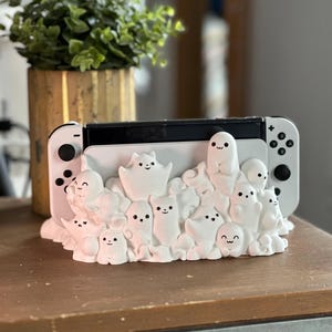 May include: White Nintendo Switch console with attached controllers, on a decorative base. The base features many small, smiling ghost figures. A potted plant is in the background, on a wooden surface.