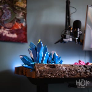 Può includere: Un gruppo di formazioni di cristalli blu con accenti gialli, montati su una base di legno scuro. I cristalli hanno un design sfaccettato e sono disposti in una forma naturale e organica. La base ha una superficie ruvida e strutturata.