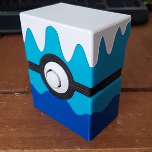May include: A 3D-printed storage box in the style of a Pok&eacute; Ball. The box is rectangular with a white top and a blue and black design. The box has a white button in the center and is sitting on a wooden surface.