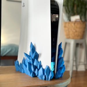 Op de afbeelding: Een witte PlayStation 5-console met een blauwe, kristalachtige basis. De console staat op een houten tafel. De blauwe kristallen zijn rond de basis van de console geplaatst.