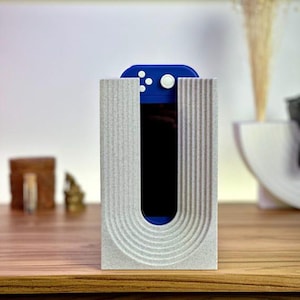 May include: A light gray, speckled, rectangular stand with a U-shaped cutout featuring vertical ribbed textures. A blue handheld gaming console with white buttons is inserted into the center of the stand, resting vertically within the cutout.
