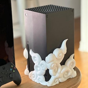 May include: A matte black rectangular gaming console featuring a grid-patterned top vent and a white, three-dimensional decorative attachment with swirling cloud-like patterns secured to the lower front and side panels of the device.