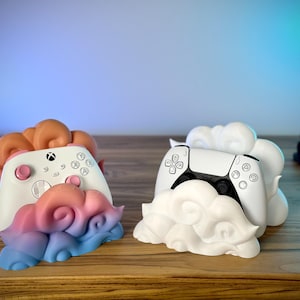 Pode incluir: Dois suportes para comandos, um para Xbox e outro para PlayStation 5. O comando Xbox repousa sobre um suporte com um design de nuvem em tons de rosa, azul e laranja. O comando PlayStation 5 repousa sobre um suporte branco em forma de nuvem.