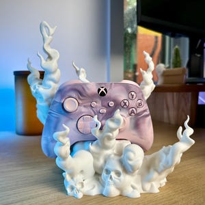 Peut inclure: Une manette Xbox personnalisée aux tons violets et roses, posée sur une base blanche en forme de crâne et de fumée. La manette affiche le logo Xbox et les étiquettes des boutons. La base présente un motif de crâne et de fumée.
