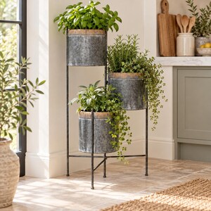 May include: A three-tier metal plant stand featuring cylindrical galvanised steel pots with ribbed copper-toned rims. The stand has a slender black iron frame with a tripod base, holding three separate containers filled with various green herbs.