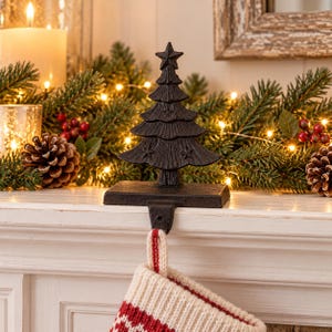 May include: A black cast iron stocking holder featuring a stylized Christmas tree design with a star at the top. The holder has a flat rectangular base and a hook extending downward to support a white and red knit stocking.