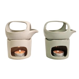 Könnte beinhalten: Zwei Keramik-Duftlampen, eine in Off-White und die andere in Hellgrün, jeweils mit einer darin brennenden Kerze. Der obere Teil hat einen Griff und einen Ausguss. Der untere Teil hat eine Öffnung für die Kerze.