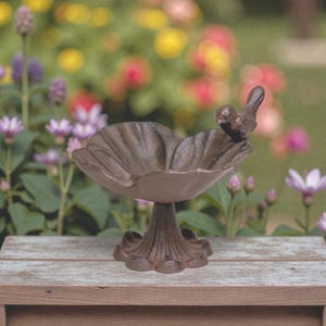 May include: A brown cast iron bird bath with a small bird perched on the edge. The bird bath has a petal-shaped bowl and a decorative pedestal base. The background is a blurred view of colorful flowers.