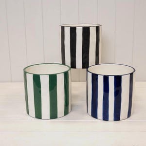 May include: Three cylindrical ceramic planters with vertical stripes in black and white, green and white, and blue and white. The planters are of different heights and stand on a white surface. The planters are suitable for indoor or outdoor use.