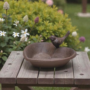 May include: A brown bird bath with a small bird statue perched on the edge. The bird bath is made of a textured material and sits on a wooden table. The background features green foliage and white flowers.