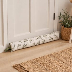 Puede incluir: Un tope de puerta cilíndrico y alargado con una base de tela color blanco hueso y un patrón repetitivo de hojas y tallos verdes. El artículo incluye una pequeña presilla de tela verde en un extremo y está colocado horizontalmente en la base de una puerta.