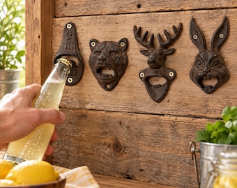 May include: Four wall-mounted cast iron bottle openers attached to a wooden board. The designs include a standard opener with the text OPEN HERE, a bear head, a deer head with antlers, and a rabbit head. One opener is in use with a glass bottle.