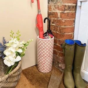 May include: A red umbrella and a black umbrella are in a patterned umbrella stand. A pair of green rain boots with blue interiors are next to the stand. A woven basket with white flowers and a wooden boot jack are also in the image.