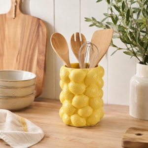 Könnte beinhalten: Ein leuchtend gelber Utensilienhalter aus Keramik mit einer strukturierten Prägung aus zahlreichen Zitronen auf der Außenseite. Der zylindrische Behälter enthält einen Holzlöffel, eine Holzgabel, einen Schneebesen aus Metall und einen Holzspatel.