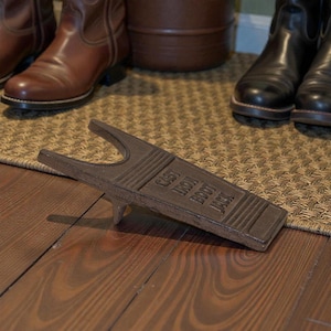 Peut inclure: Un tire-bottes en fonte de couleur marron avec l'inscription "CAST IRON BOOT JACK" en relief. Le tire-bottes est posé sur un sol en bois. Plusieurs paires de bottes sont en arrière-plan.