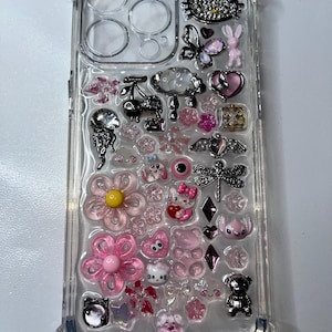 May include: Clear phone case decorated with various pink and silver charms. Charms include flowers, hearts, a Hello Kitty, a teddy bear, a dragonfly, and a butterfly. The case is made of clear plastic.
