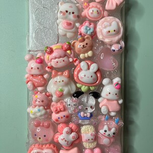 May include: A clear phone case adorned with an assortment of pink and white kawaii-style charms. The charms include various animals, bows, hearts, and stars. The case is designed to protect the phone while adding a cute, decorative element.