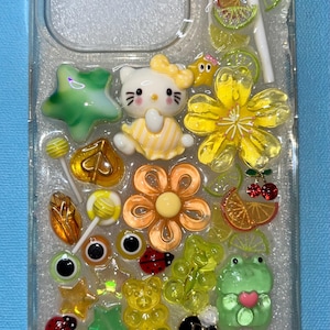 Op de afbeelding: Een transparante telefoonhoes met een wit Hello Kitty-personage, groene en gele sterren, bloemen en andere kleurrijke charmes. De hoes is gevuld met glitter en heeft een witte achtergrond.