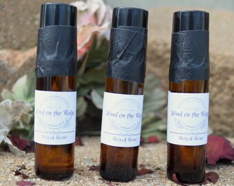May include: Three amber glass bottles with black roll-on tops and white labels. The labels have a purple floral design and the text "Wind on the Ridge" and "Royal Rose".