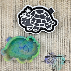 May include: Two turtle-shaped craft molds. One is black and white, the other is a translucent green. Both have a smiling face and a hat. The molds are on a textured, light brown surface. The bottom right corner has the text "Pine & Feathers".