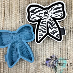 May include: A black and white zebra print bow-shaped accessory, with a matching blue silicone mold. The bow has a black border and a black ribbon. The bow is a craft supply for making hair accessories or decorations.