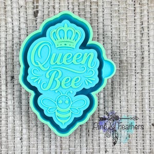 May include: A teal and blue cookie cutter with the words "Queen Bee" and a bee graphic. The design includes a crown above the text and is surrounded by a scalloped edge. The cutter is made of plastic.