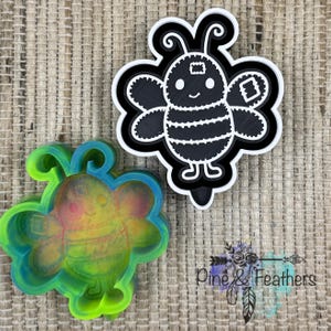 May include: Two bee-shaped cookie cutters. One is a black and white design, and the other is a translucent green, blue, and yellow. The cutters are on a textured, neutral-colored surface. The black and white cutter has a detailed bee design.