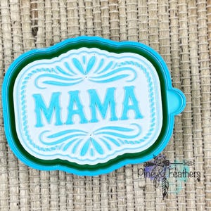 May include: A turquoise and white cookie cutter with the word "MAMA" in a decorative font. The cutter has a rectangular shape with ornate floral designs and a turquoise border.