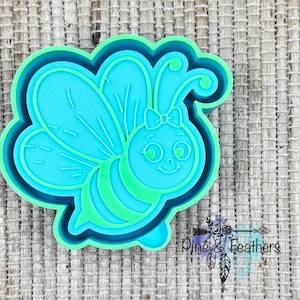 May include: A turquoise and green bee-shaped cookie cutter. The bee has large wings, a smiling face, and a bow. The cutter is made of plastic and has a detailed design. The image is of a cookie cutter, likely for baking.