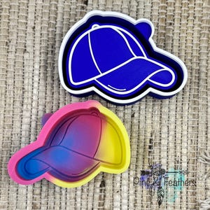 May include: Two baseball cap-shaped cookie cutters. One is blue with white accents, and the other is a gradient of pink, blue, and yellow. The cutters are made of plastic and are designed for baking or crafting. The background is a woven, neutral-toned surface.