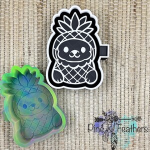 May include: A black and white pineapple-shaped cookie cutter with a cute face and a handle. A second, iridescent pineapple-shaped mold is also visible. The image also includes the text "Pine & Feathers".