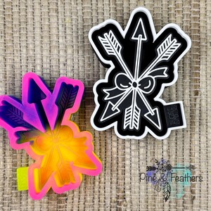 May include: Two arrow-themed cookie cutters. One is a black and white plastic cutter featuring three crossed arrows tied with a bow. The other is a vibrant, multi-colored gradient plastic cutter in shades of pink, purple, orange, and yellow.