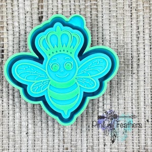 May include: A turquoise and green bee-shaped cookie cutter with a crown. The bee has a smiling face, wings, and striped body. The cutter is layered with a dark blue outline and a light green outline. The text "Pine & Feathers" is in the bottom right corner.
