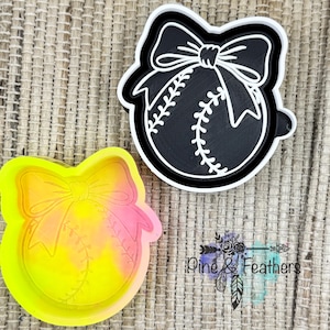 May include: Two baseball-themed cookie cutters. One is black and white, featuring a baseball with a bow. The other is a tie-dye of yellow and pink. The cutters are on a textured, neutral surface. The logo "Pine & Feathers" is visible.