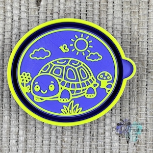 May include: A blue and yellow turtle-themed cookie cutter. The design features a cartoon turtle, sun, clouds, and flowers. The cookie cutter is made of plastic and has a rounded shape.