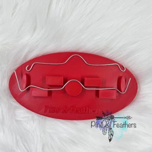 May include: A red oval-shaped plastic base featuring a silver wire structure attached to the top. The base has several small rectangular and circular raised supports and the text Pine & Feathers embossed on the front surface.