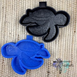 May include: A black sea turtle cookie cutter and a blue sea turtle cookie cutter. Each cutter features a detailed shell pattern and a protruding handle tab at the top. The cutters are shaped like swimming sea turtles with textured flippers.