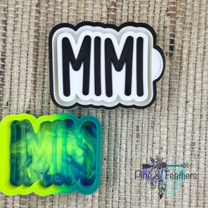 May include: Two MIMI-shaped cookie cutters. One is black, white, and gray, and the other is green and blue. The MIMI text is in bold, black letters. The cookie cutters are on a textured, tan surface.