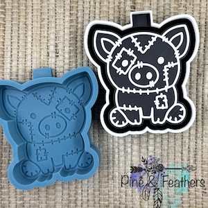 May include: A blue silicone mold and a black and white pig-shaped design. The pig has stitched patches and a cute expression. The design is outlined in white and black. The mold is a light blue color. The design has the text "Pine & Feathers".