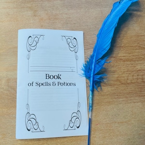 Può includere: Un libro bianco vuoto con il titolo "Book of Spells & Potions" e bordi decorativi. Una penna stilografica a penna blu giace su una superficie di legno accanto al libro.