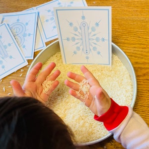 Può includere: Le mani di un bambino tengono riso bianco in una ciotola d'argento. Sullo sfondo ci sono delle carte con fiocchi di neve blu su una superficie di legno.